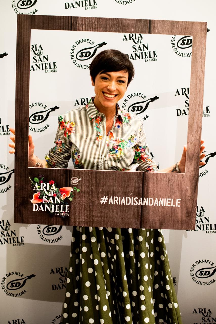 Rubina Rovini posa con la cornice di Aria di San Daniele davanti al fondale del Consorzio del Prosciutto di San Daniele.