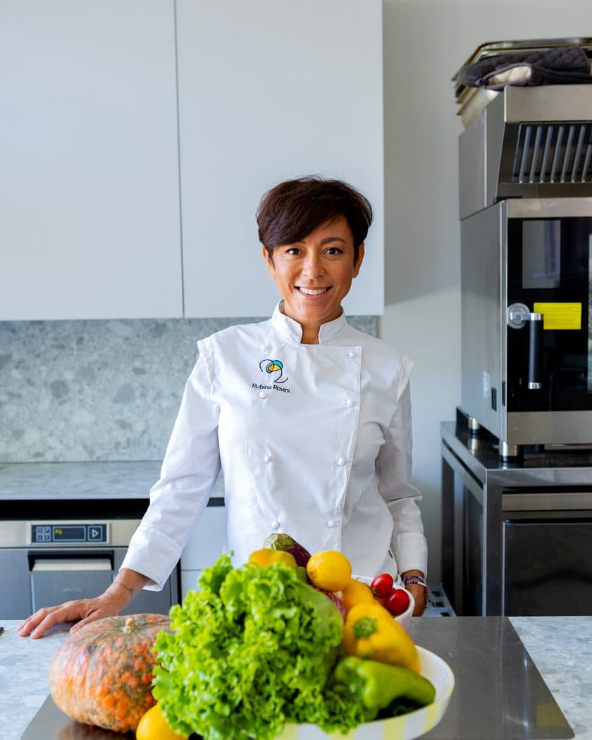 Rubina Rovini in giacca da chef dietro al banco di RADICI Lab, con verdure di stagione.