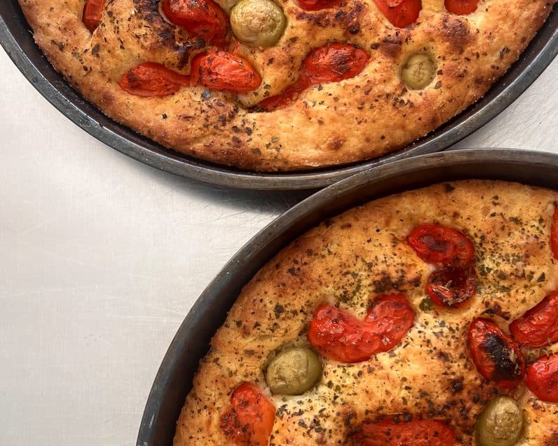 Due focacce con pomodorini e olive appena sfornate nelle teglie di ferro.