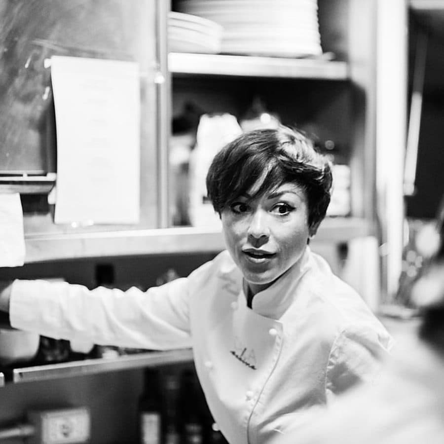 Rubina Rovini in giacca da chef al passe della cucina, fra le mensole del servizio.