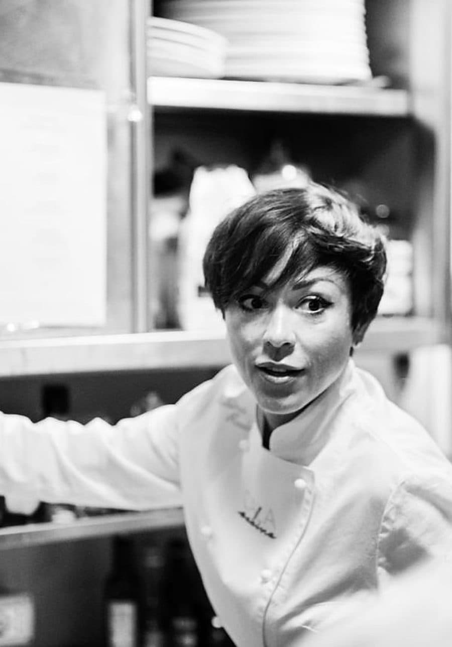 Rubina Rovini in giacca da chef si volta verso l'obiettivo dal passe di una cucina professionale.
