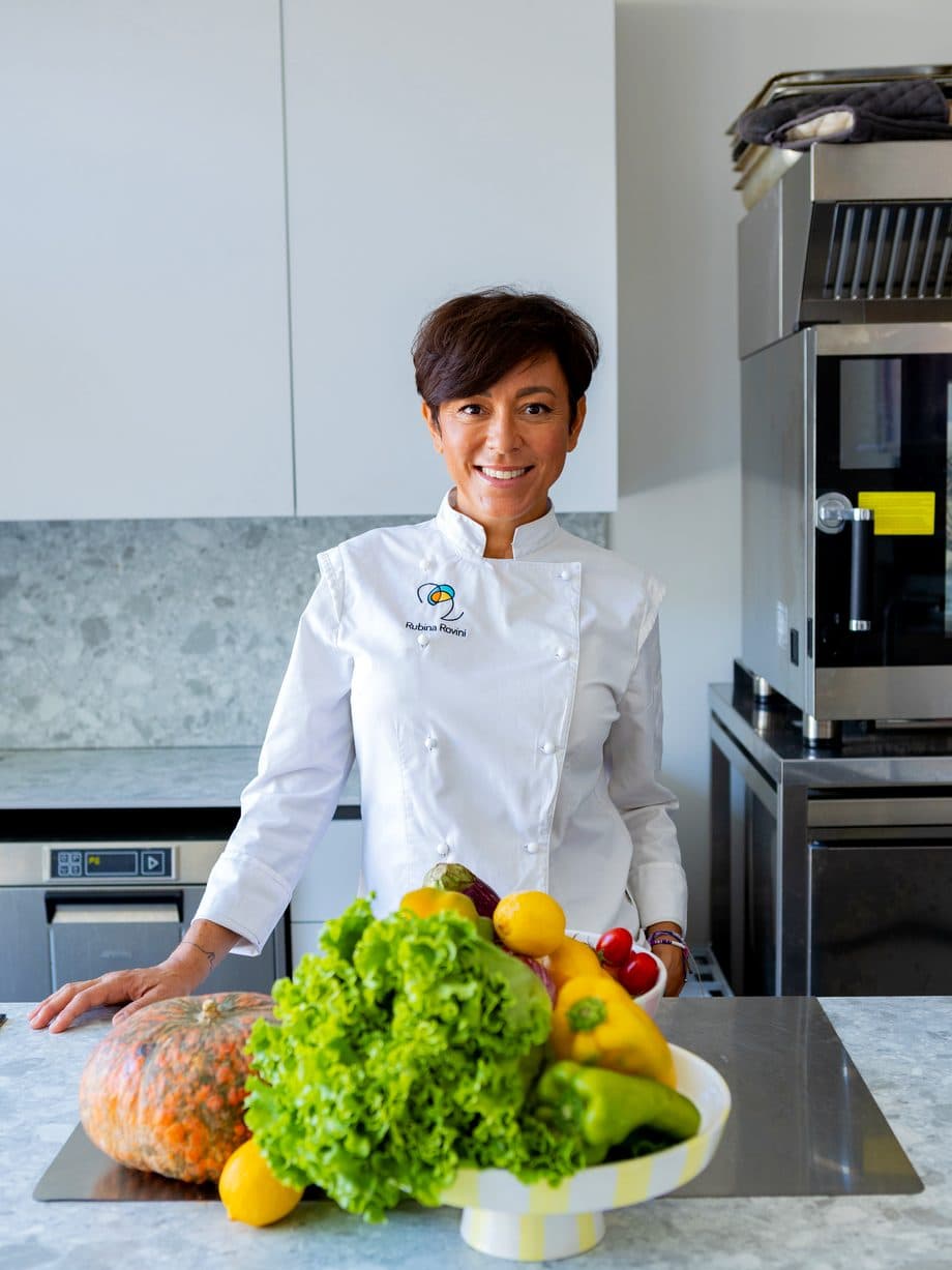 Rubina Rovini in giacca da chef dietro al banco di RADICI Lab, con verdure di stagione.
